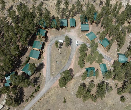 Aerial view of a wooded area with several green-roofed cabins arranged along a circular dirt road, with two parked vehicles and scattered trees throughout.