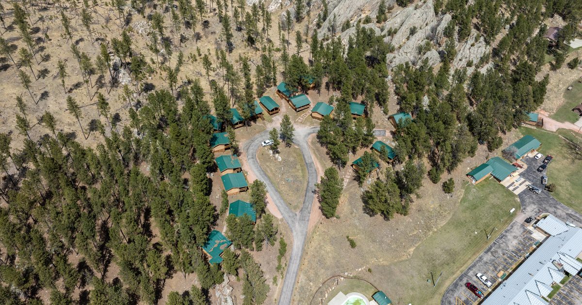 Aerial view of a group of green-roofed cabins arranged in a semicircle among pine trees near rocky hills, with a road, a parking lot, and an outdoor pool visible on the right side.