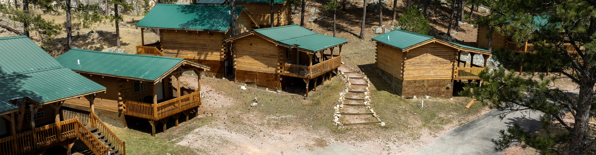 A group of wooden cabins with green roofs nestled among tall pine trees, connected by dirt paths and steps, with a paved road curving in front of them.