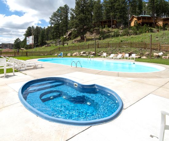 Wide shot of the outdoor, in-ground hot tub with the pool in the background. Past the pool area is a hill with cabins on top of it.