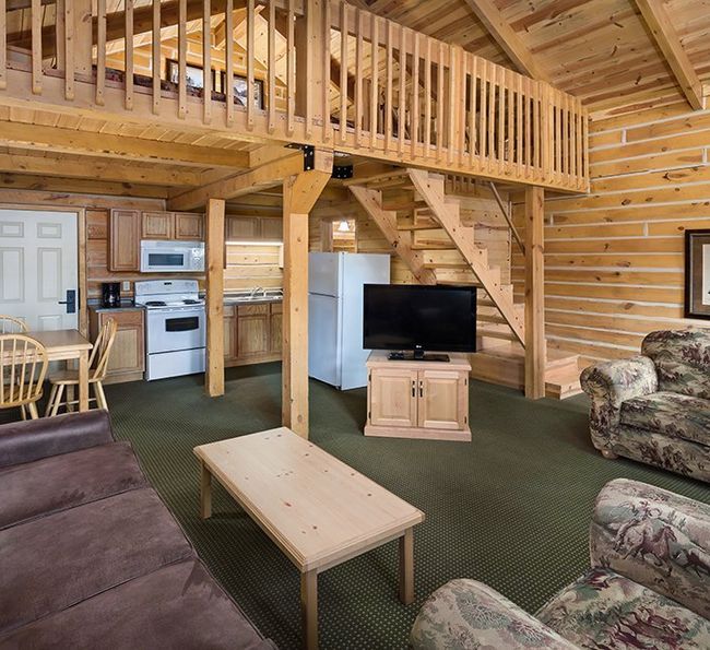 A cozy log cabin interior with wood-paneled walls, a loft, a small kitchen, a dining table, a TV, a brown sofa, and patterned armchairs. A framed picture hangs on the wall above one chair.