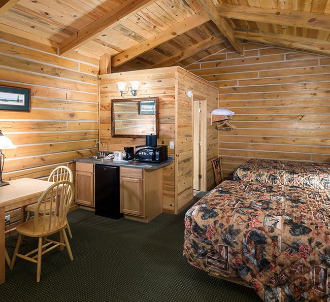 A cozy log cabin room with two beds, a small dining table with chairs, wooden walls and ceiling, a kitchenette with a microwave and coffee maker, and soft lighting from table lamps.