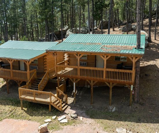 A wooden cabin with a green metal roof sits on a slope in a forested area, supported by stilts. It has two covered porches, a central staircase, and is surrounded by pine trees and rocky terrain.