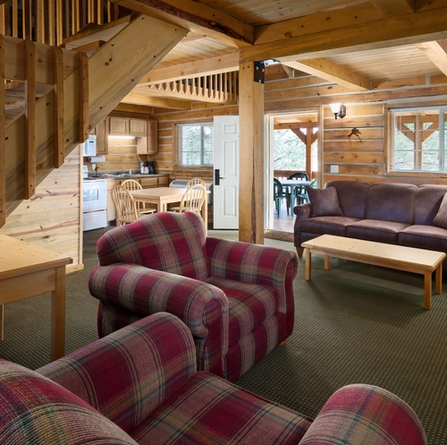 The first floor of a cabin, with the living room as the focal point. The walls have a wood paneling. There are two plaid chairs next to the staircase that leads to the second floor. There is also a brown leather couch and a TV turned on to a baseball game. In the back is part of the kitchen with a table and four wooden chairs.