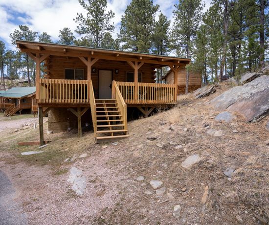 A small wooden cabin with a front porch and steps sits on a sloped, rocky hillside surrounded by pine trees. Other cabins are visible in the background under a partly cloudy sky.