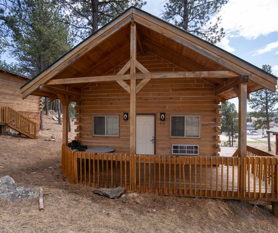 A small wooden cabin with a covered porch and railing sits on a sloped, pine-covered area. Two chairs and a table are on the porch. Other similar cabins are visible nearby under a partly cloudy sky.