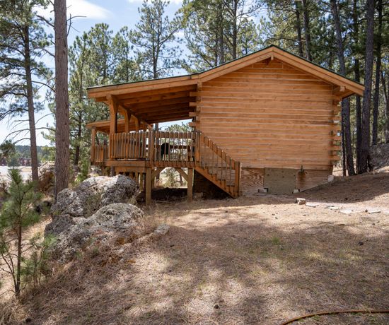 Side angle of the exterior of a cabin. It has log siding and a deck that overlooks the piny, rocky terrain.