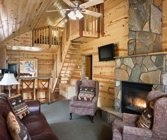 A cozy log cabin living room with wood-paneled walls, a stone fireplace, leather sofa, patterned armchairs, a loft area with wooden stairs, and a small kitchen with bar stools in the background.