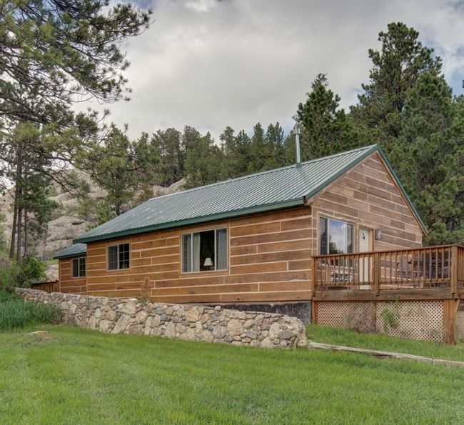 A wooden cabin with a green metal roof and a spacious deck sits surrounded by pine trees and grass, with a stone retaining wall in front and partly cloudy skies above.