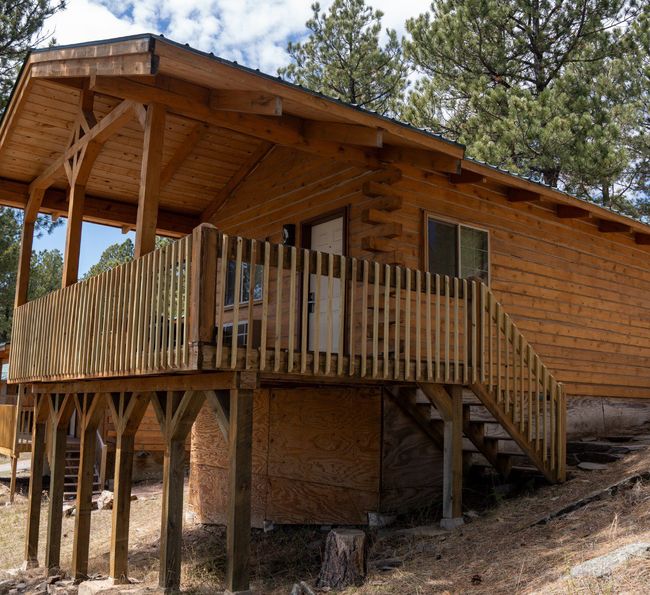 A wooden cabin stands on stilts in a forested area, with a front porch, railing, and stairs leading up to the entrance. Pine trees and a partly cloudy sky are visible in the background.
