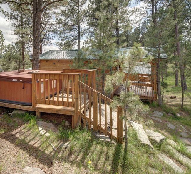 A wooden cabin with a green metal roof is nestled among tall pine trees. A red hot tub sits on a raised deck with wooden railings, surrounded by green grass and rocks in a peaceful, forested area.