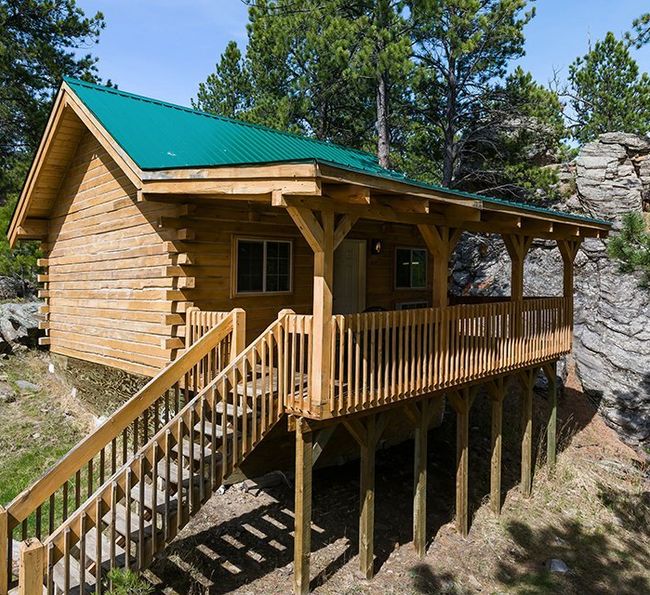 A small wooden cabin with a green metal roof and a raised porch, surrounded by pine trees and rocks, set on a hillside with stairs leading up to the entrance.