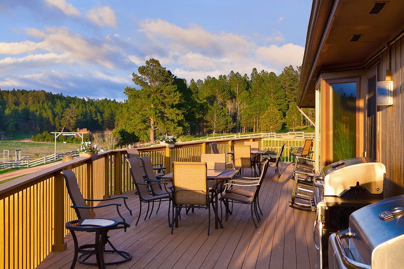 A spacious wooden deck with several tables and chairs overlooks green fields and pine trees at sunset. Gas grills are on the right, and flower boxes line the railing. The sky is partly cloudy.