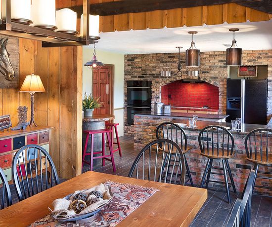 A rustic kitchen and dining area with wood and brick accents, black spindle chairs, a wooden dining table, a kitchen island with barstools, pendant and chandelier lighting, and horse-themed decor on the wood-paneled wall.