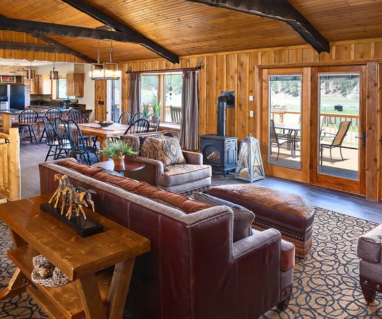 A cozy rustic living room with brown leather sofas, a wood-burning stove, wooden walls and ceiling, large windows and doors, and an open view into a dining area and kitchen in the background.