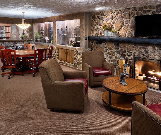 Cozy living room with stone walls, a lit fireplace, a large TV above the mantel, and comfortable brown chairs around a wooden coffee table. A dining table with red chairs sits near a built-in aquarium on the left.