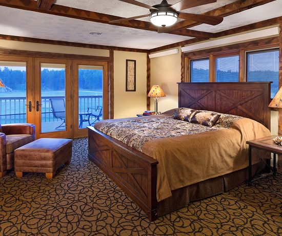 A cozy bedroom with a large wooden bed, patterned bedding, two matching lamps, an armchair with an ottoman, carpeted floor, and glass doors opening to a balcony with outdoor seating and a scenic view.