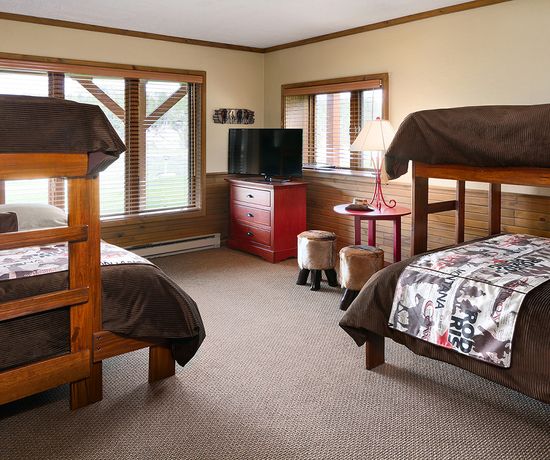 A cozy bedroom with two wooden bunk beds, a red dresser with a TV, a lamp on a side table, and windows letting in natural light. The room has brown décor and beige carpeting.