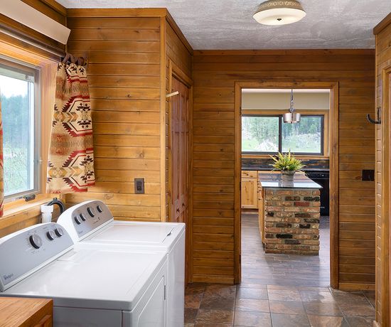 A laundry room with a washer and dryer under a window with patterned curtains; a wooden doorway leads to a kitchen with wood cabinets and a brick island.