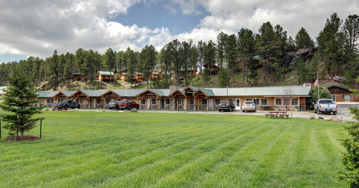 Single-story cabins with green roofs line the edge of a large, neatly mowed grassy field, surrounded by pine trees and small hills under a partly cloudy sky. Several cars and picnic tables are visible near the buildings.