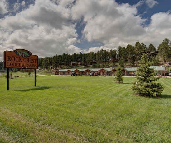 The front of the property. The sign is on the left side, and it says "Rock Crest Lodge & Cabins". The lodge is in the background. There are cars parked in front of it. In the background is a small hill where the cabins sit.