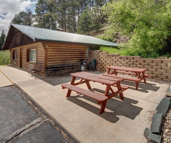 A log-cabin style building built partially into a hill. It sits on the pavement with two red wooden picnic tables next to it.