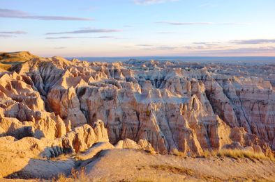 Eroded rock formations and rugged cliffs in warm sunlight at Badlands National Park, with a vast, layered landscape extending to the horizon under a partly cloudy sky.