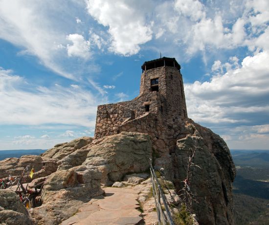A stone lookout tower stands atop a rocky mountain peak under a partly cloudy sky, with distant forests and hills visible in the background.
