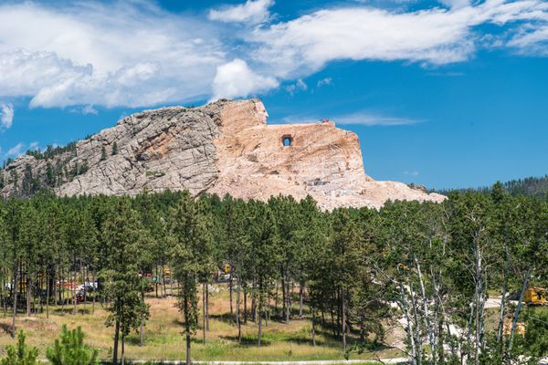 Crazy Horse Monument from far away. His face is carved from the rock and there is a hole beneath it that shows the blue sky beyond. A pine forest lies beneath the monument.