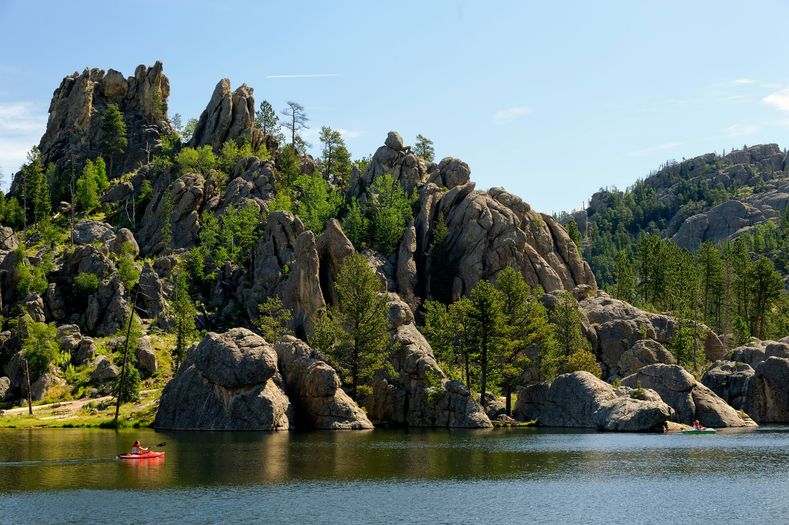 Rocky cliffs with green pine trees rise above a calm lake, where two people in colorful kayaks paddle near the shore under a clear blue sky.