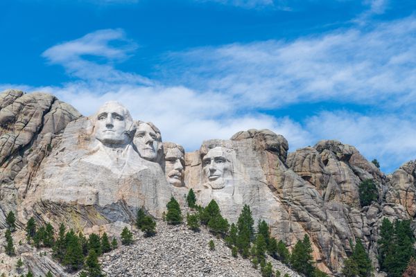 Mount Rushmore. Four former presidents' faces are carved into the rock. There is blue sky surrounding it and pine trees standing beneath it.