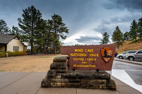 A rustic sign that says "Wind Cave National Park Visitor Center and Headquarters". A building sits on the left-hand side and the parking lot with a couple cars parked in it on the right. The sky is gray and there are pine trees near the building and parking lot.
