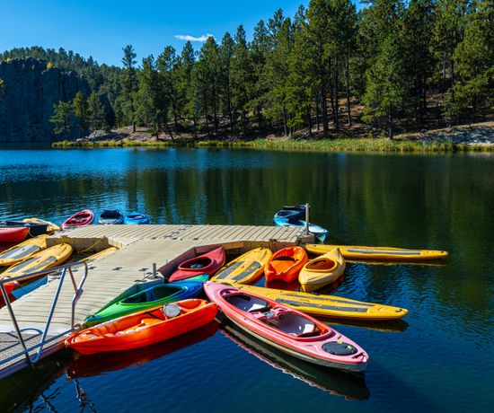 Colorful kayaks and canoes are lined up on a wooden dock floating on a calm lake surrounded by pine trees and rocky cliffs under a clear blue sky.