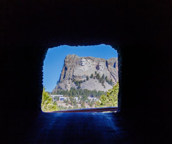 View of Mount Rushmore framed by the dark interior of a tunnel, with blue sky and green trees visible outside, highlighting the monument’s carved presidential faces in the distance.