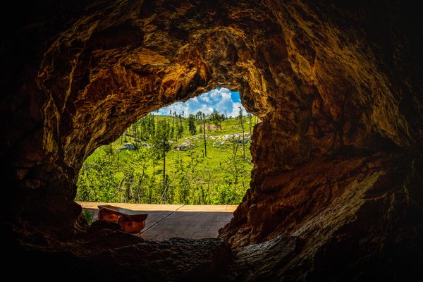 View from inside a rocky cave looking out onto a sunny, green landscape with trees and blue sky. A wooden bench sits at the cave entrance on a stone path.